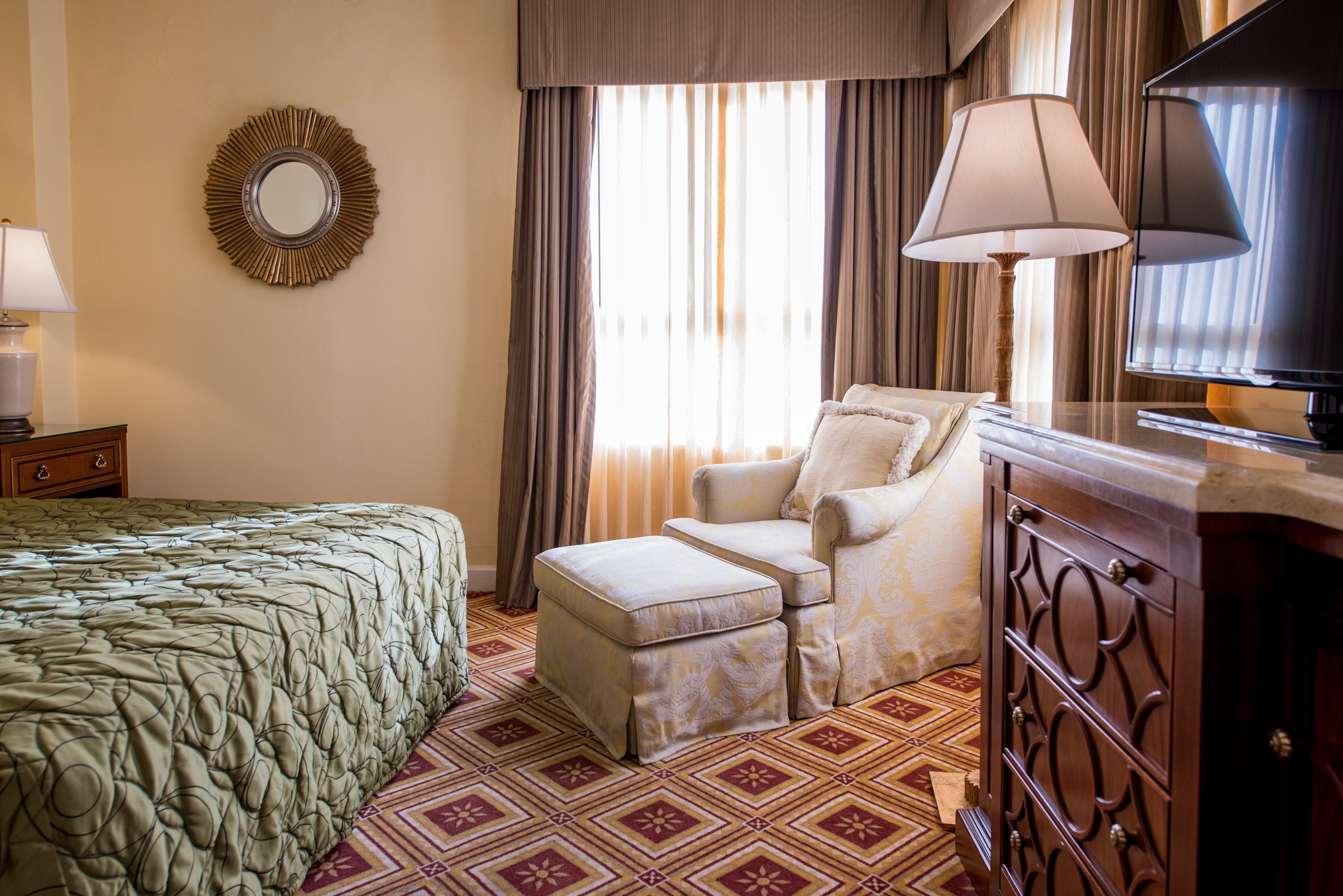 A hotel room with a bed, a chair, a TV, a lamp, and a mirror on the wall.