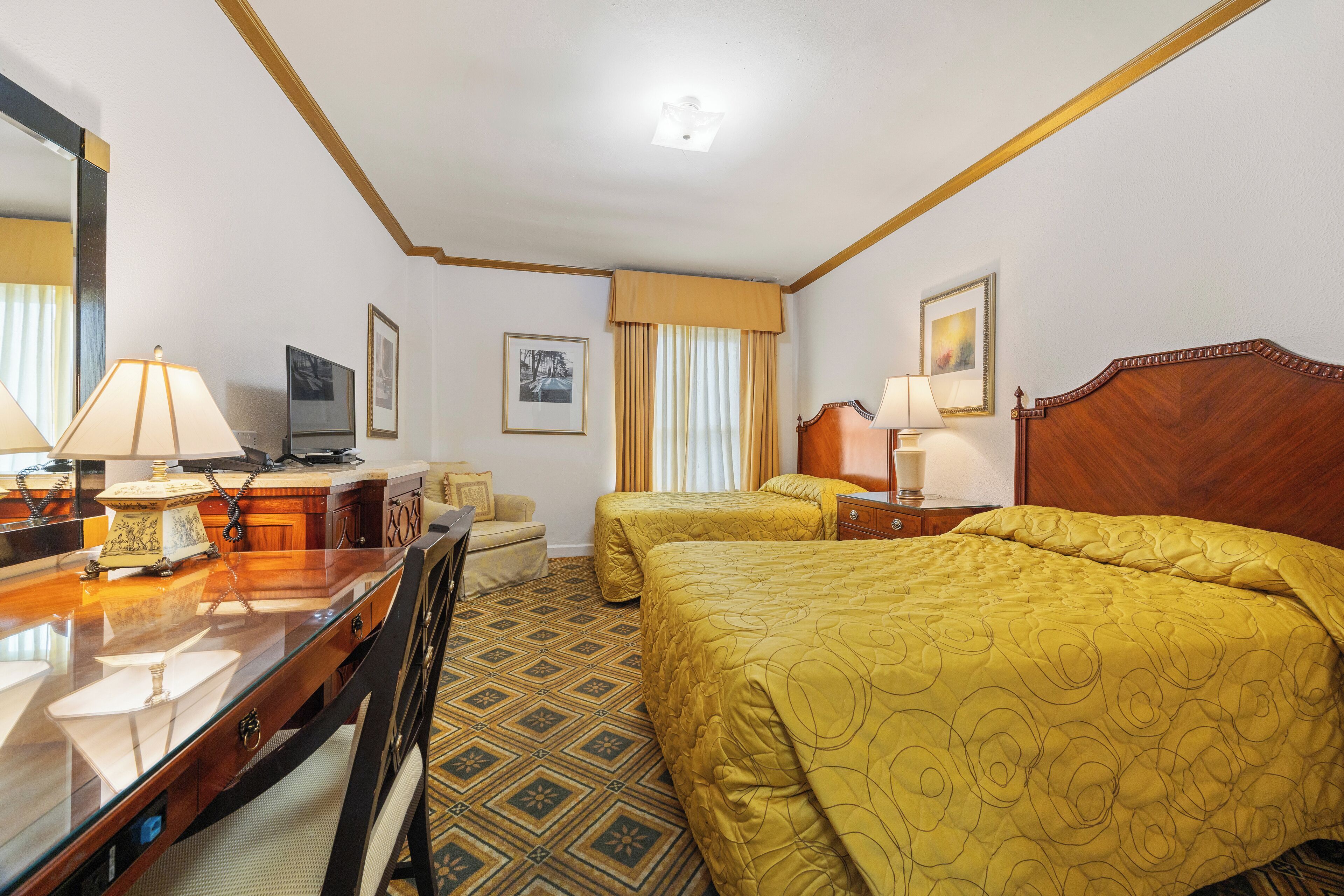 A hotel room with a wooden desk, two beds with yellow bedding, a large mirror, and a television.