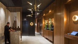 Un área de recepción moderna con una recepcionista en un escritorio de vidrio, un mostrador de madera, una planta decorativa y una pantalla digital.
