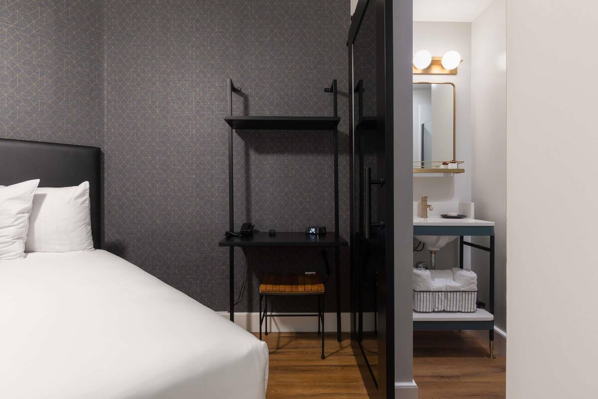 A hotel room with a bed, a desk with a chair, a vanity with a mirror, and a towel rack.