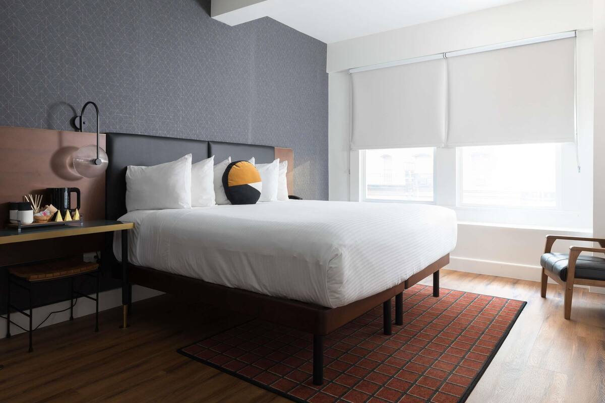 A modern hotel room with a large bed, a desk with a lamp, and a view of the outdoors through the window.