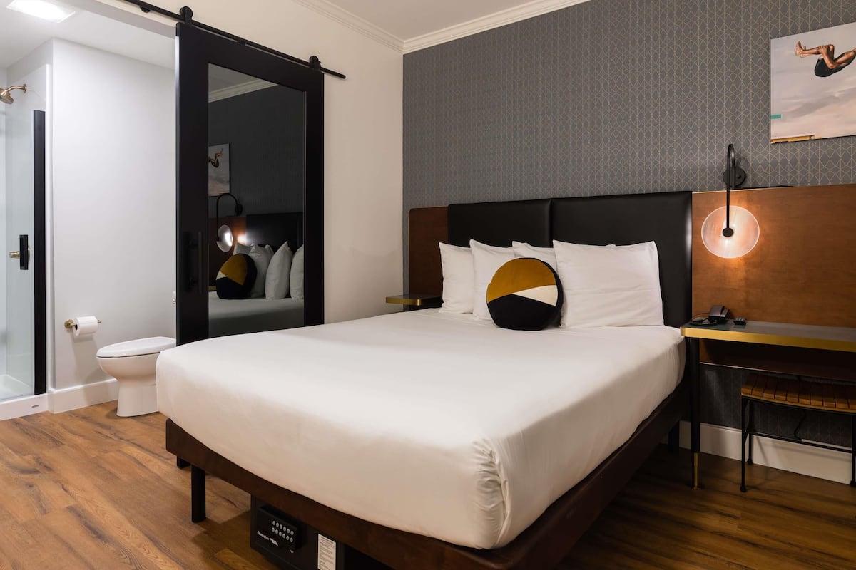 A hotel room with a large bed, a bedside table, a nightstand, a wall-mounted lamp, and a mirror.