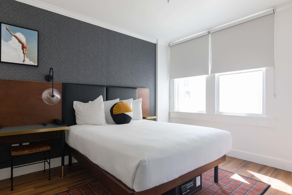 A modern hotel room with a large bed, a desk with a chair, a standing lamp, and a window with blinds.