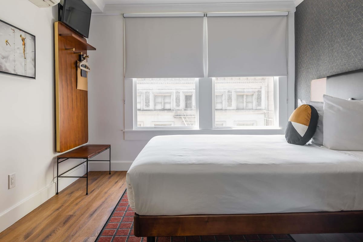 A modern hotel room with a large bed, a wooden bench, a TV, and a window with a view of buildings.