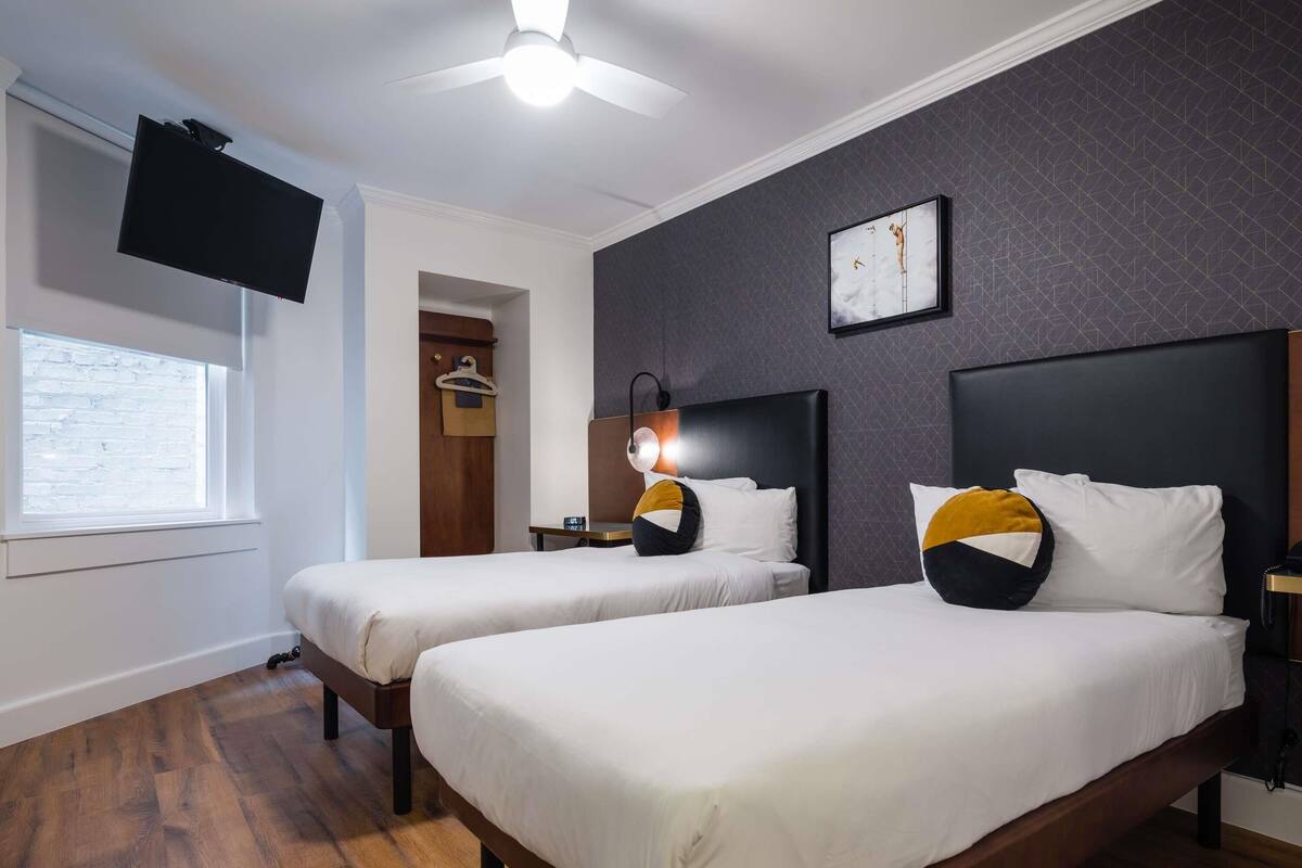 Une chambre d’hôtel avec deux lits, un ventilateur de plafond, un téléviseur fixé au mur, une horloge et une fenêtre donnant sur un mur en briques.
