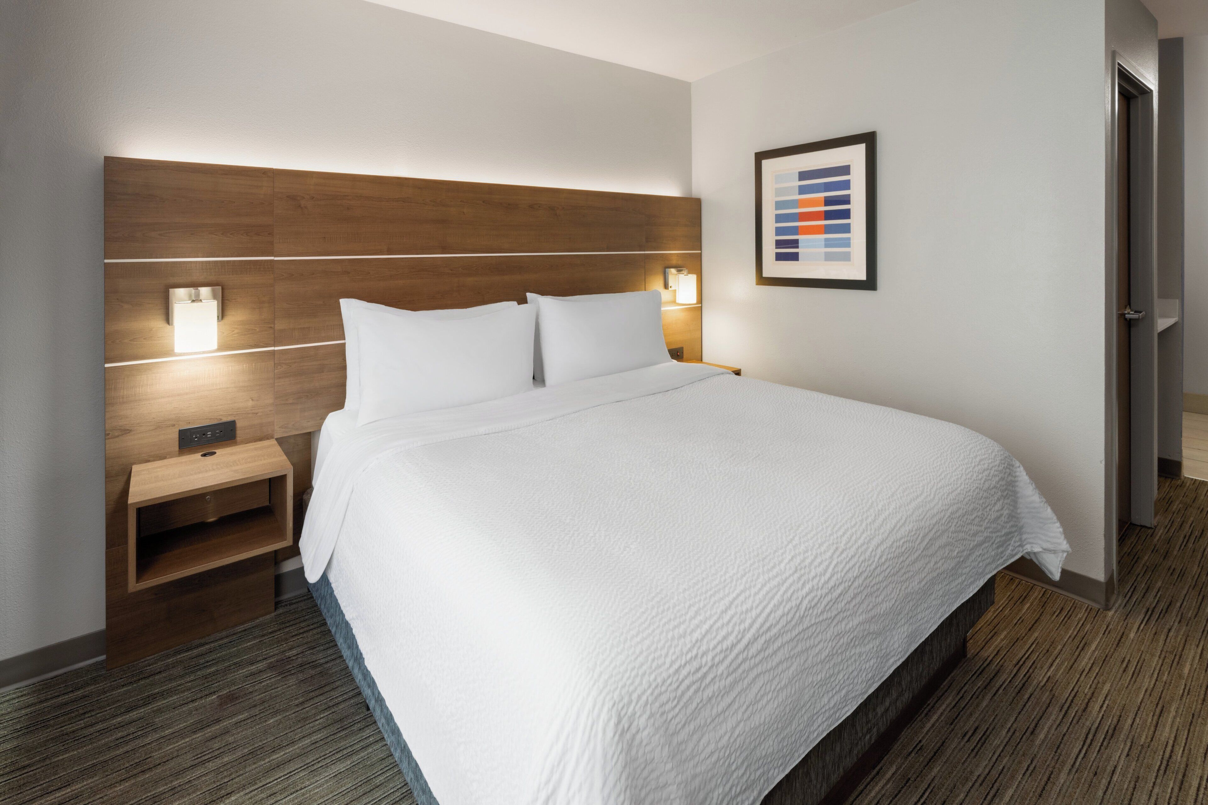 Ein Hotelzimmer mit einem großen Bett, Nachttischen, einer Wandleuchte und einem gerahmten Bild.