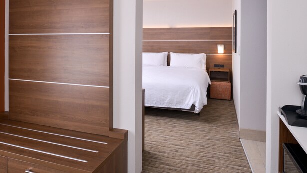 Habitación de hotel con cabecera de madera, cama con ropa blanca, mesita de noche con lámpara y una pequeña zona de estar con un banco.