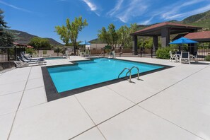 Una piscina rectangular con borde negro, rodeada por una terraza con baldosas. Se observan sillas de descanso y una zona de estar cubierta con una sombrilla azul.