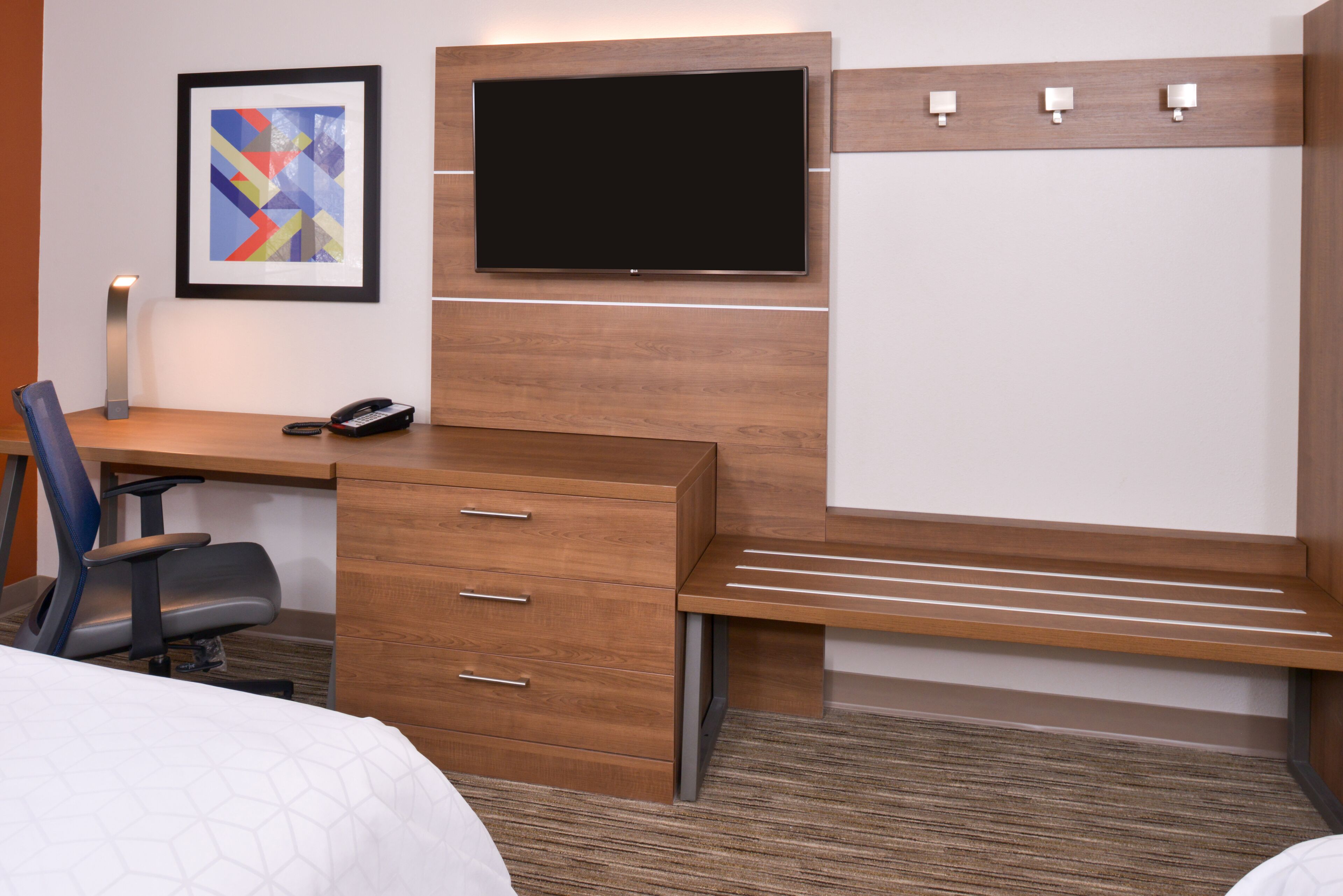 Habitación de hotel con escritorio de madera, una silla, una cama, televisor montado en la pared y un cuadro abstracto enmarcado.