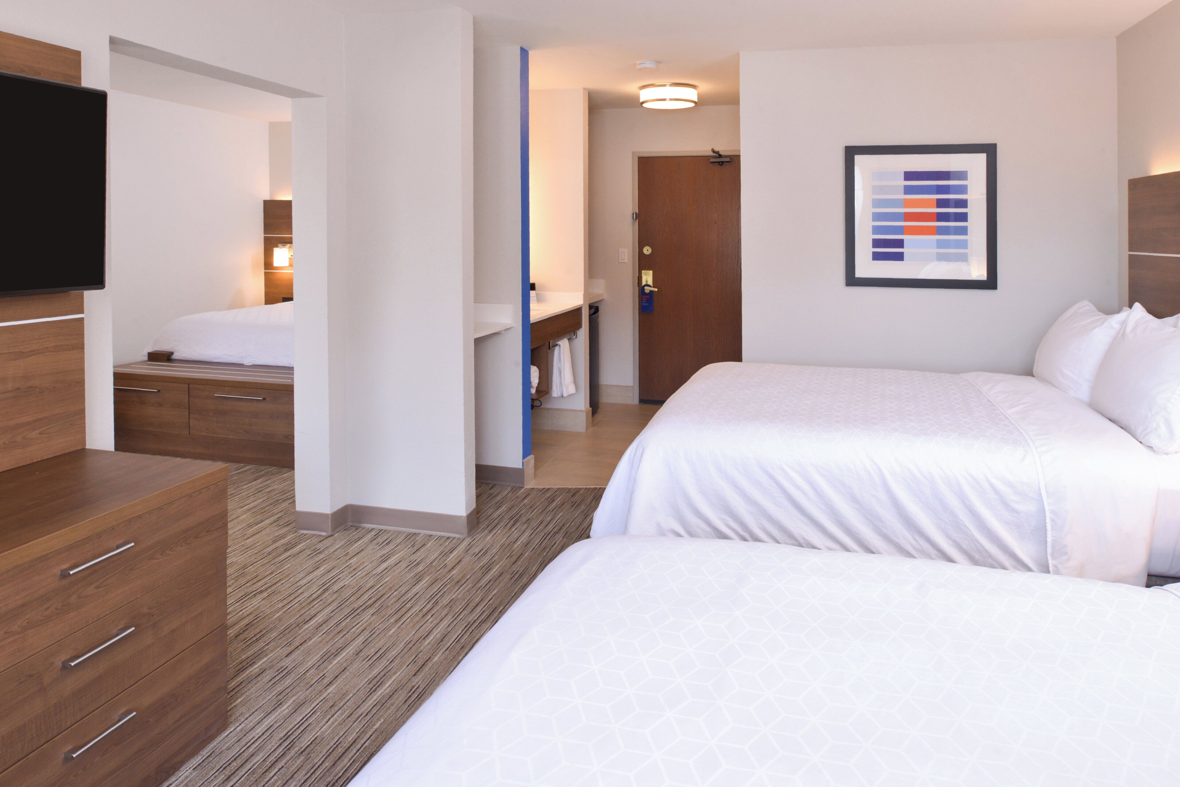 Una habitación de hotel con cama, tocador, televisor y baño visible a través de una puerta abierta.
