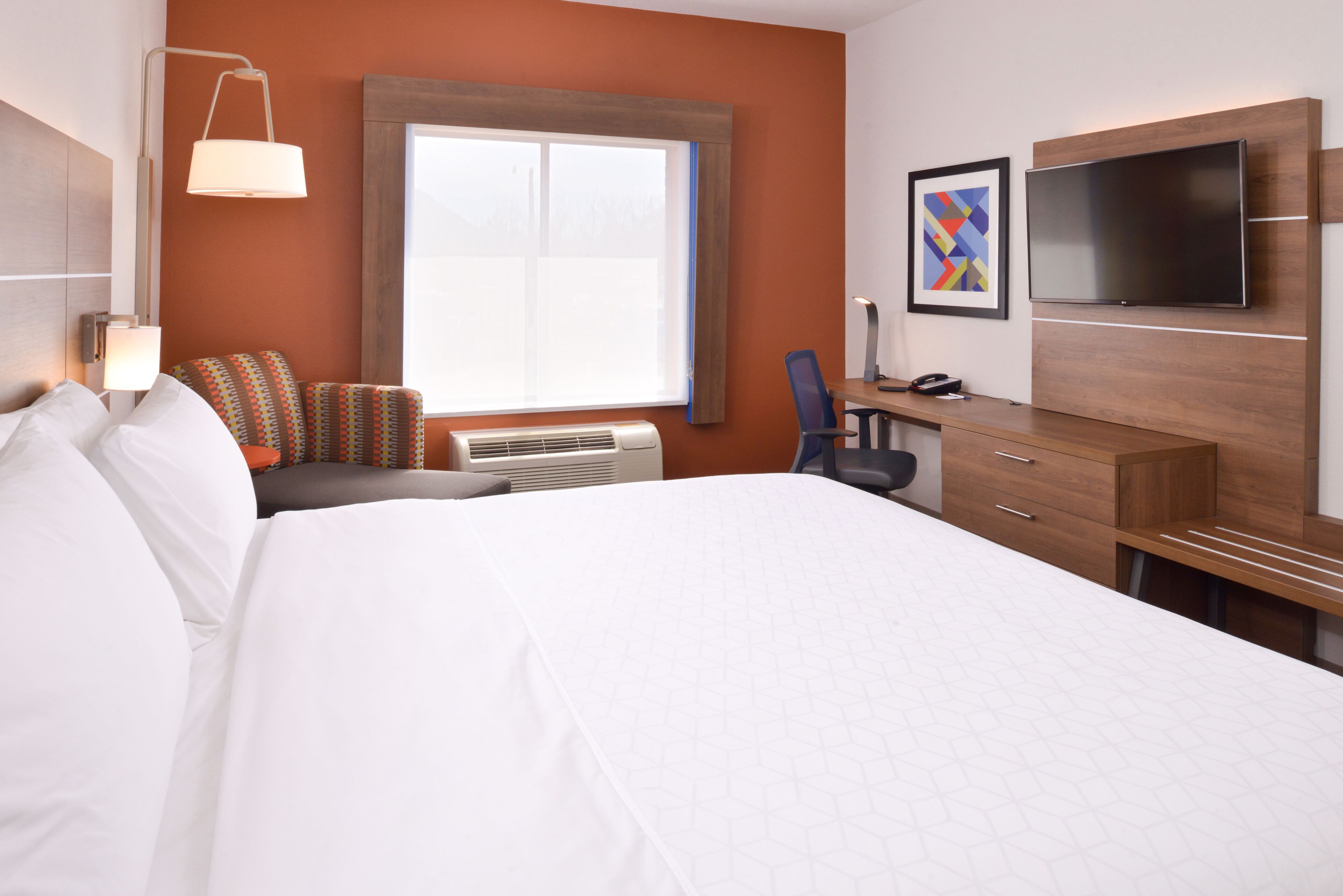 Une chambre d’hôtel dotée d’un grand lit, d’un bureau avec une chaise, d’un téléviseur à écran plat, d’une fenêtre avec des stores et d’un climatiseur fixé au mur.
