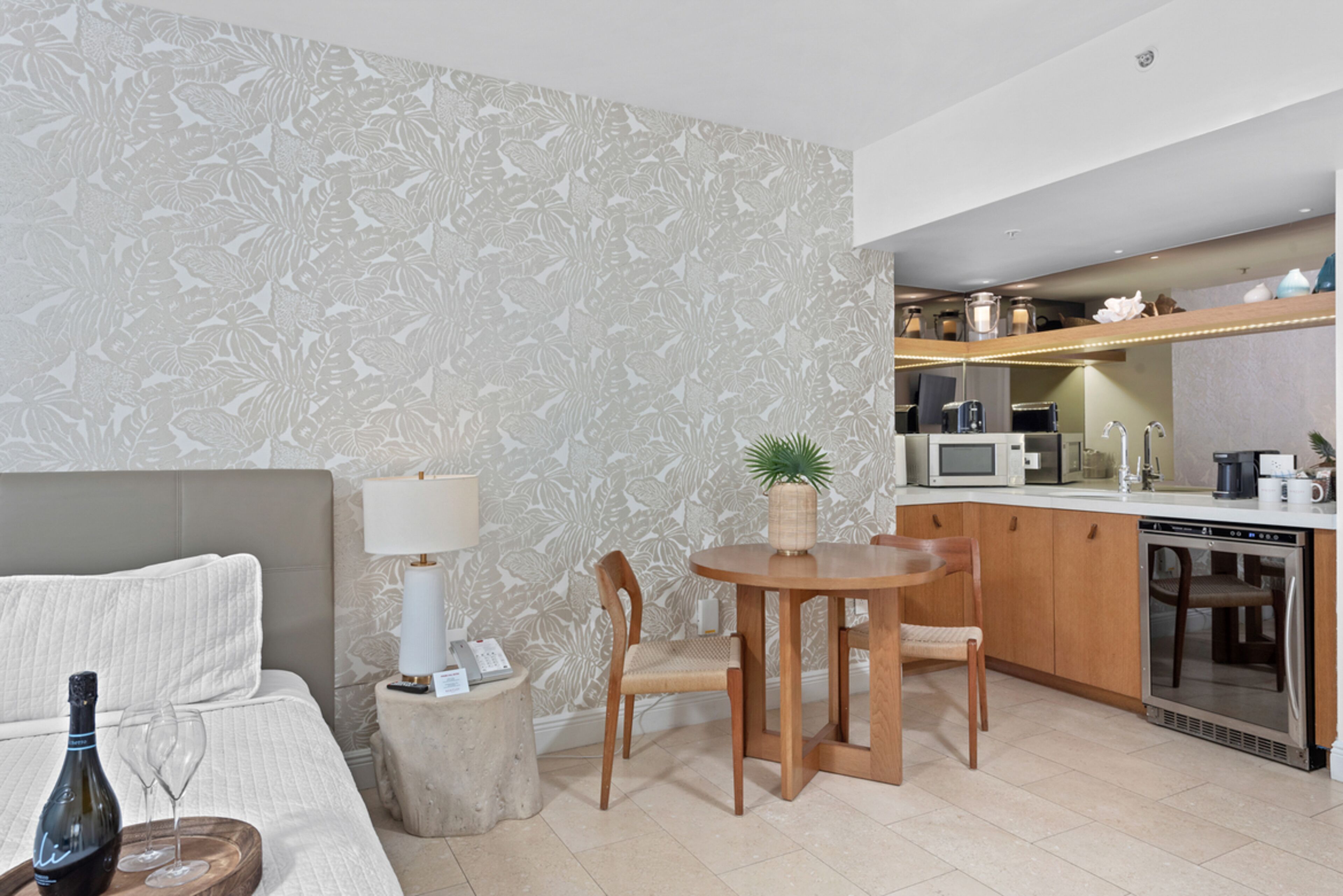 A modern hotel room with a bed, a small round table with chairs, a kitchen area with a microwave, toaster, and coffee maker, and a decorative plant.