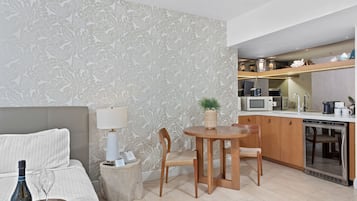 A modern hotel room with a bed, a small round table with chairs, a kitchen area with a microwave, toaster, and coffee maker, and a decorative plant.