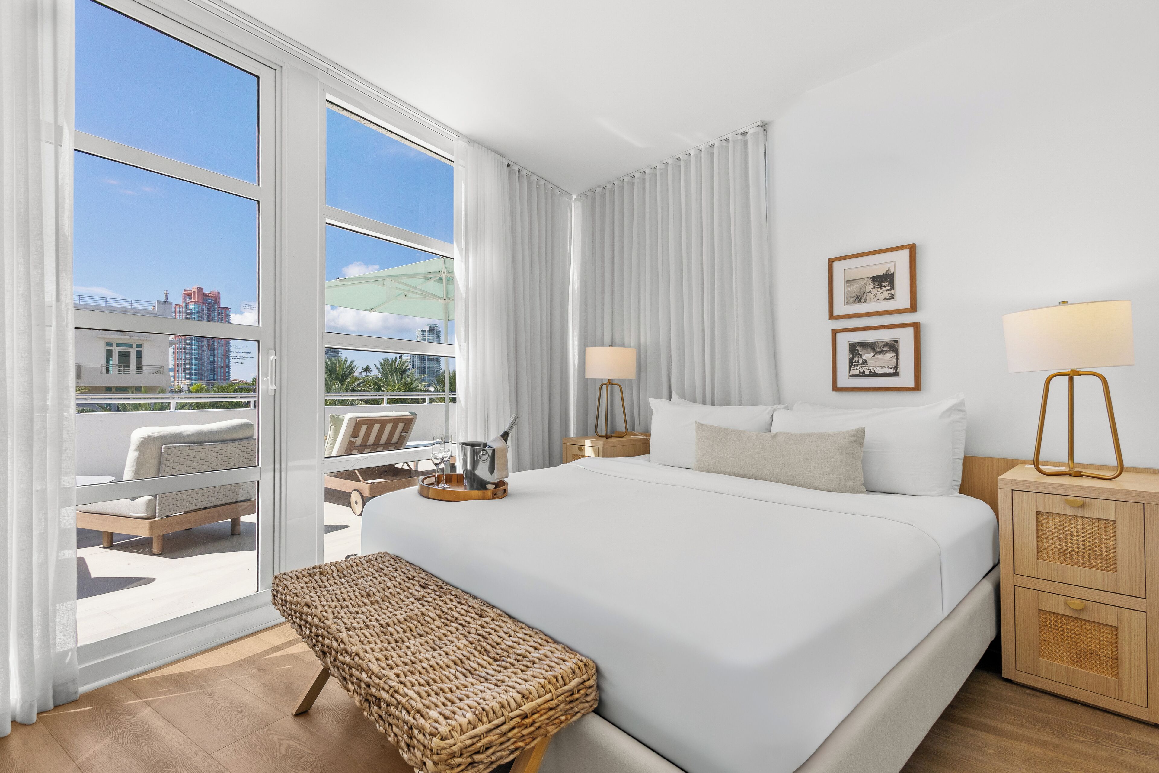 A hotel room with a large bed, a bedside table with a lamp, a bench, and a view of the city through large windows.