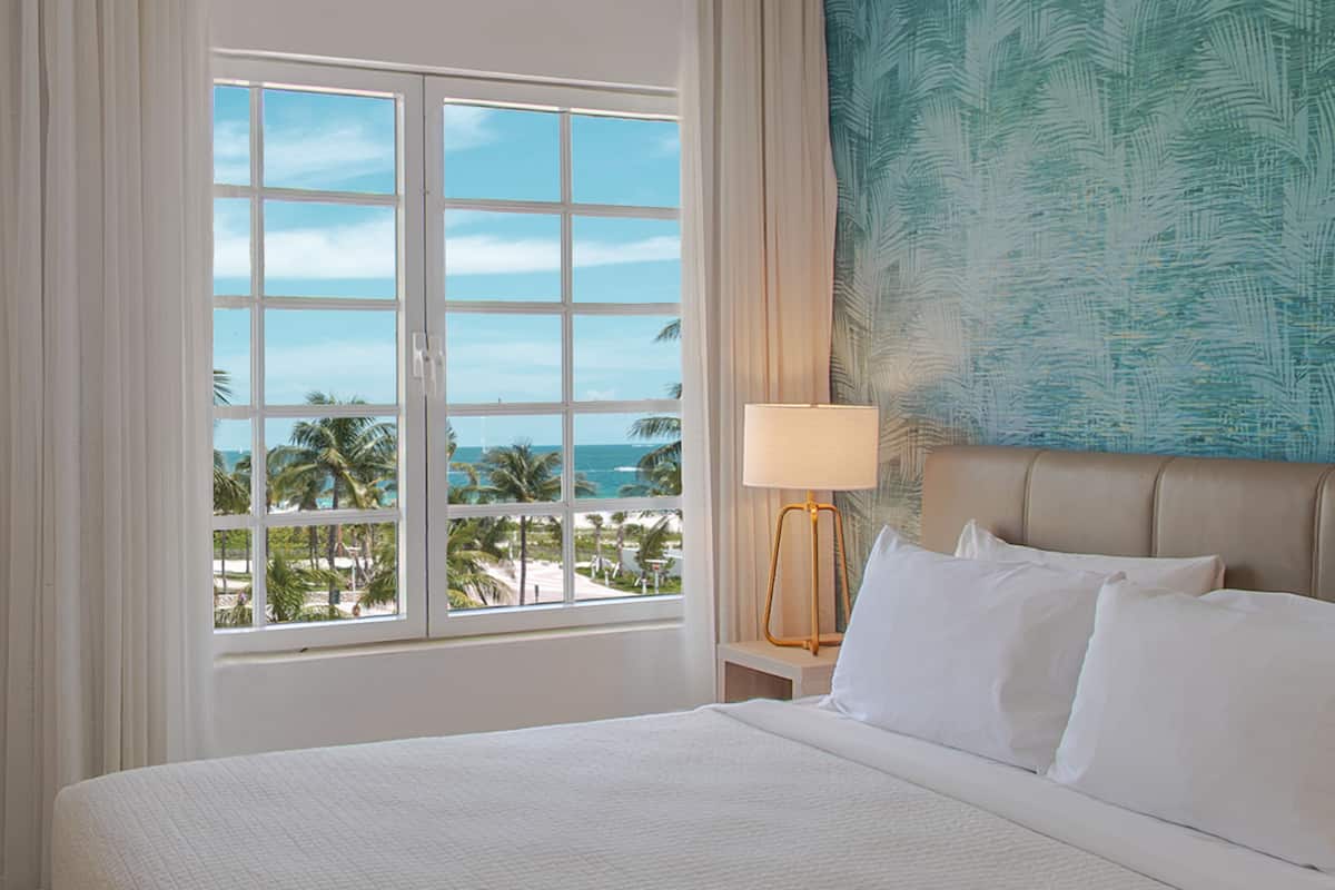 Une chambre d’hôtel dotée d’une grande fenêtre, d’un lit avec du linge de lit blanc, d’une tête de lit beige et d’un mur orné d’un motif de feuilles tropicales.