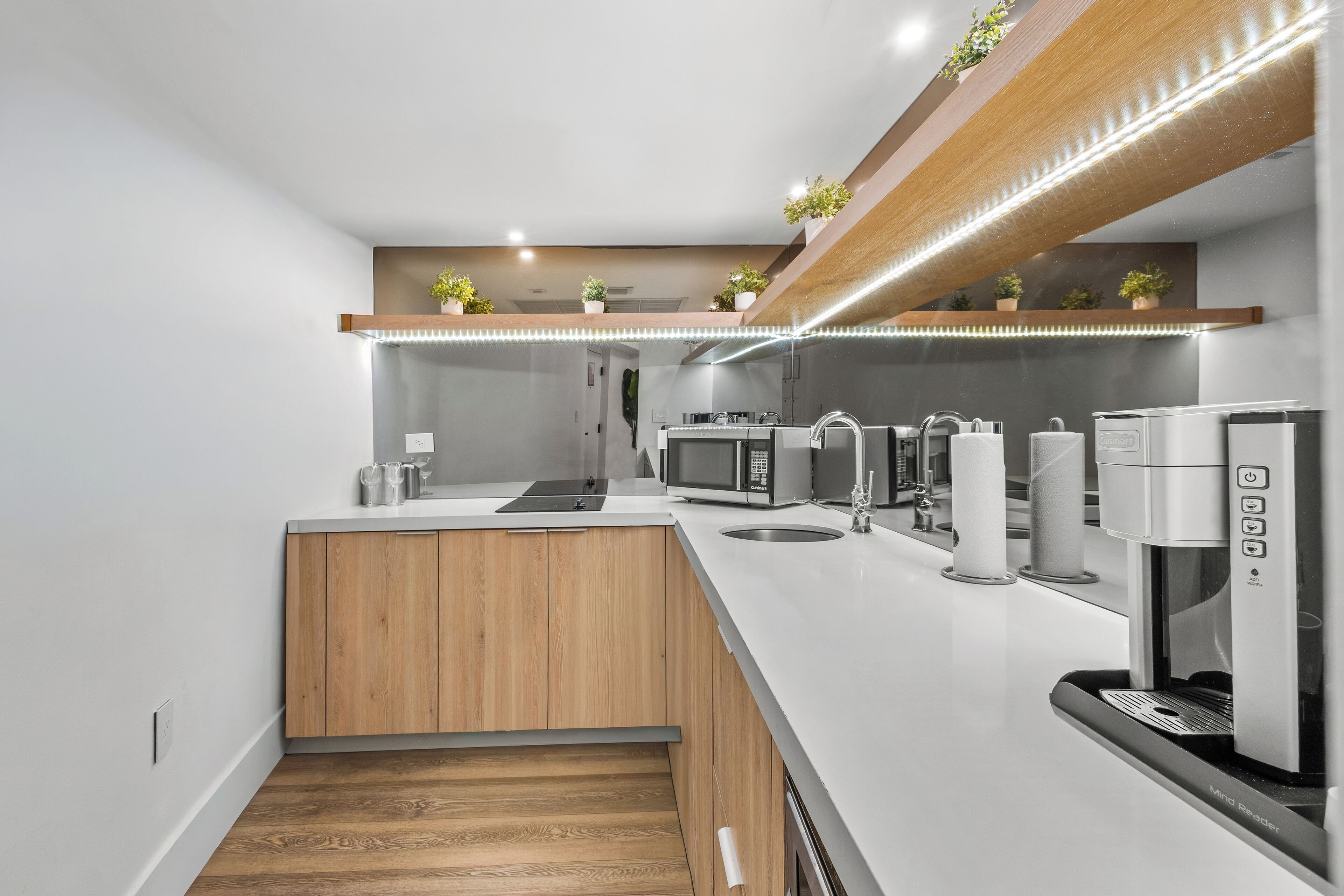 A modern kitchen with a white countertop, wooden cabinets, a built-in microwave, a coffee machine, and a small island with a sink.