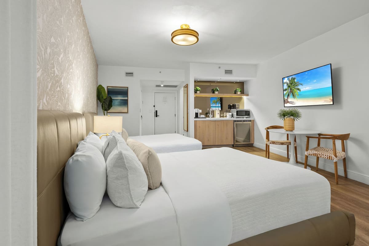 Une chambre d’hôtel avec deux lits, une kitchenette, un coin repas et un téléviseur diffusant une scène de plage.