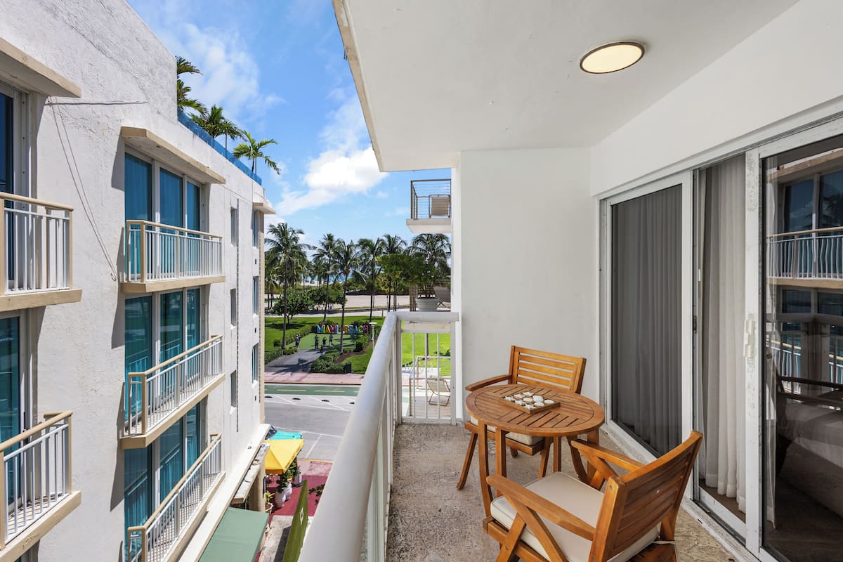 Un balcon avec une table et des chaises, une vue sur un espace verdoyant agrémenté de palmiers, et un immeuble doté de nombreuses fenêtres et de balcons.