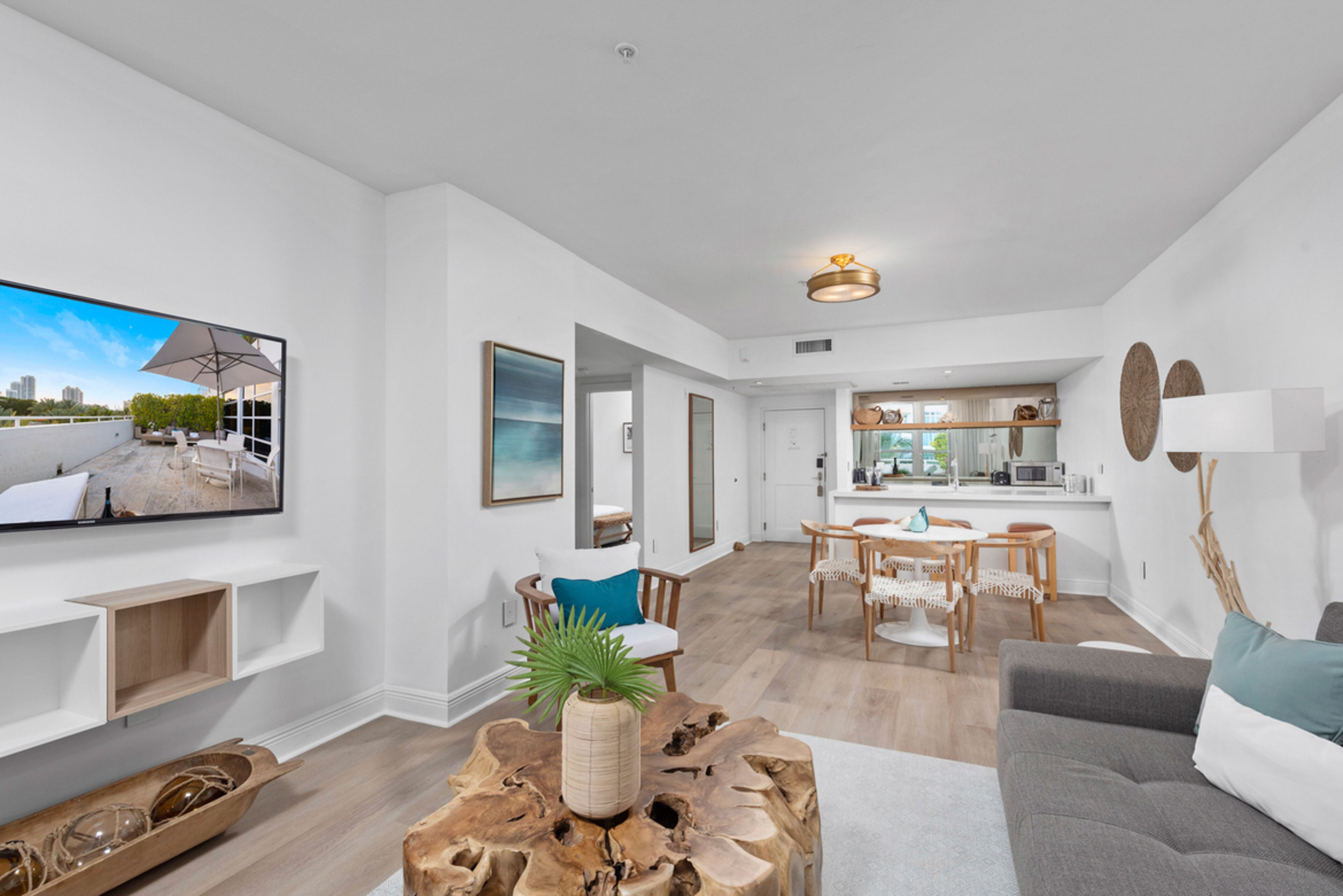 A modern living room with a flat-screen TV, wooden coffee table, and a grey sofa. There is a dining area with a wooden table and chairs.