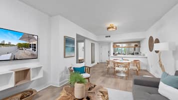 Uma sala de estar moderna com televisão de tela plana, mesa de centro de madeira e um sofá cinza. Há uma área de jantar com mesa e cadeiras de madeira.