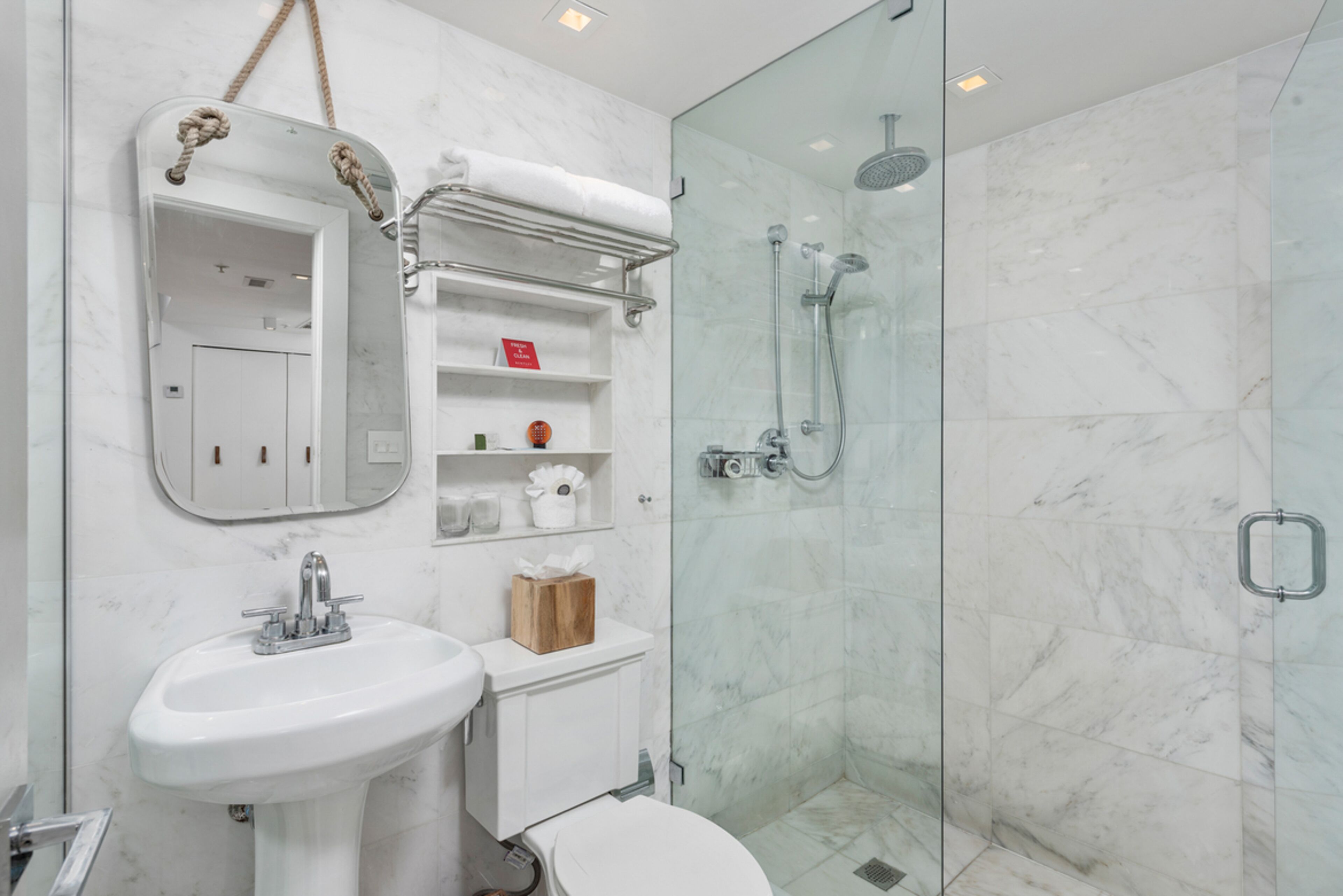 Une salle de bain moderne avec une douche à parois de verre, un lavabo blanc et un miroir doté d’une poignée en corde.