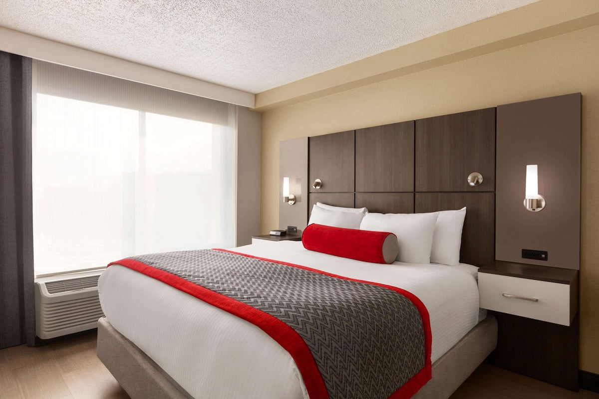 A hotel room with a large bed, a bedside table, a nightstand, a window with curtains, and a wall-mounted light fixture.