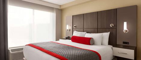 A hotel room with a large bed, a bedside table, a nightstand, a window with curtains, and a wall-mounted light fixture.