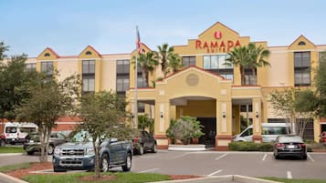 Un hotel Ramada Suites con fachada amarilla, palmeras y un estacionamiento con varios coches.