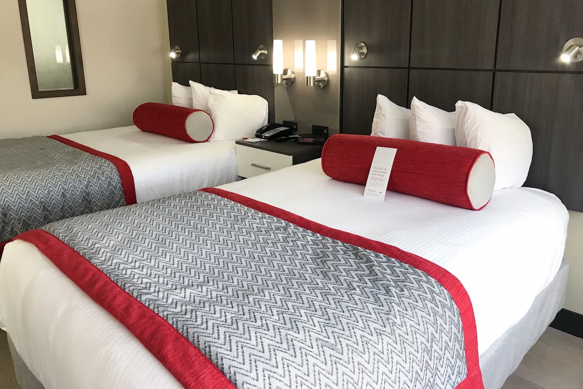 A hotel room with two beds, each with a red and white bedspread, a nightstand with a phone, and a wall-mounted light fixture.