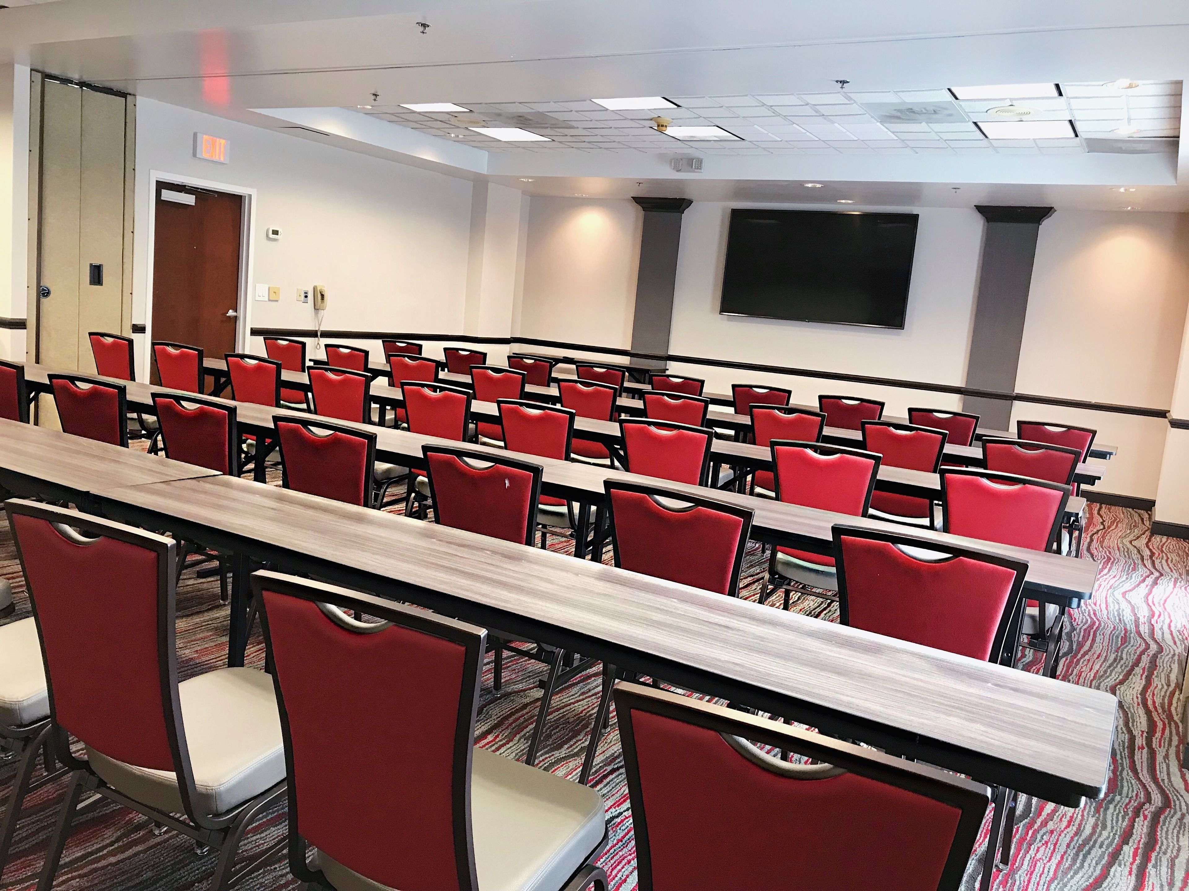 A conference room with rows of red chairs and wooden tables, a large flat-screen TV on the wall, and an exit sign above the door.