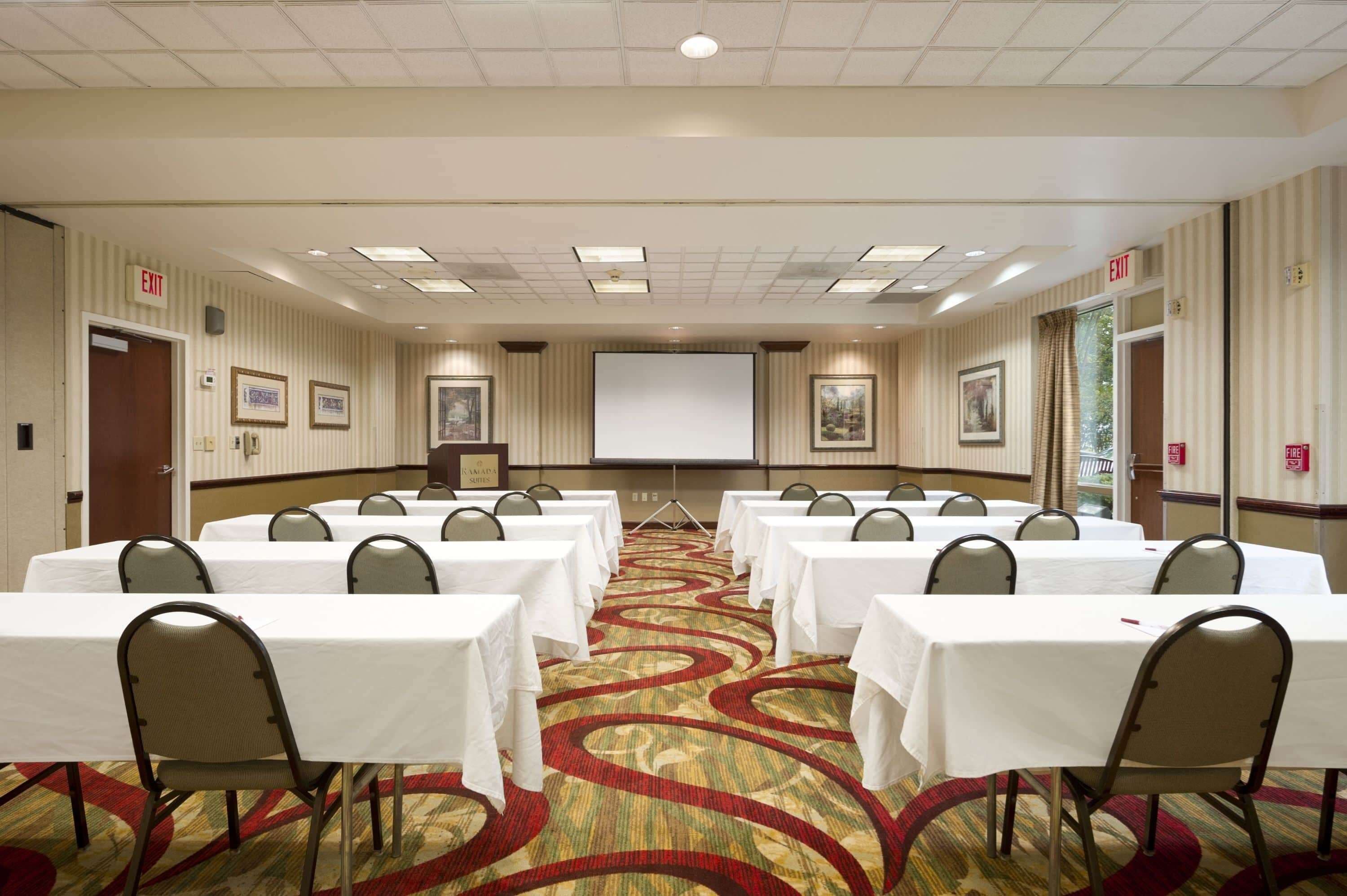 Une salle de conférence équipée de plusieurs tables et chaises, d’un écran de projection et d’un panneau indiquant la sortie.