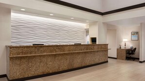 A reception desk with a textured wall behind it, a small desk with a lamp, and a chair. - Ramada by Wyndham Suites Orlando Airport (Orlando)