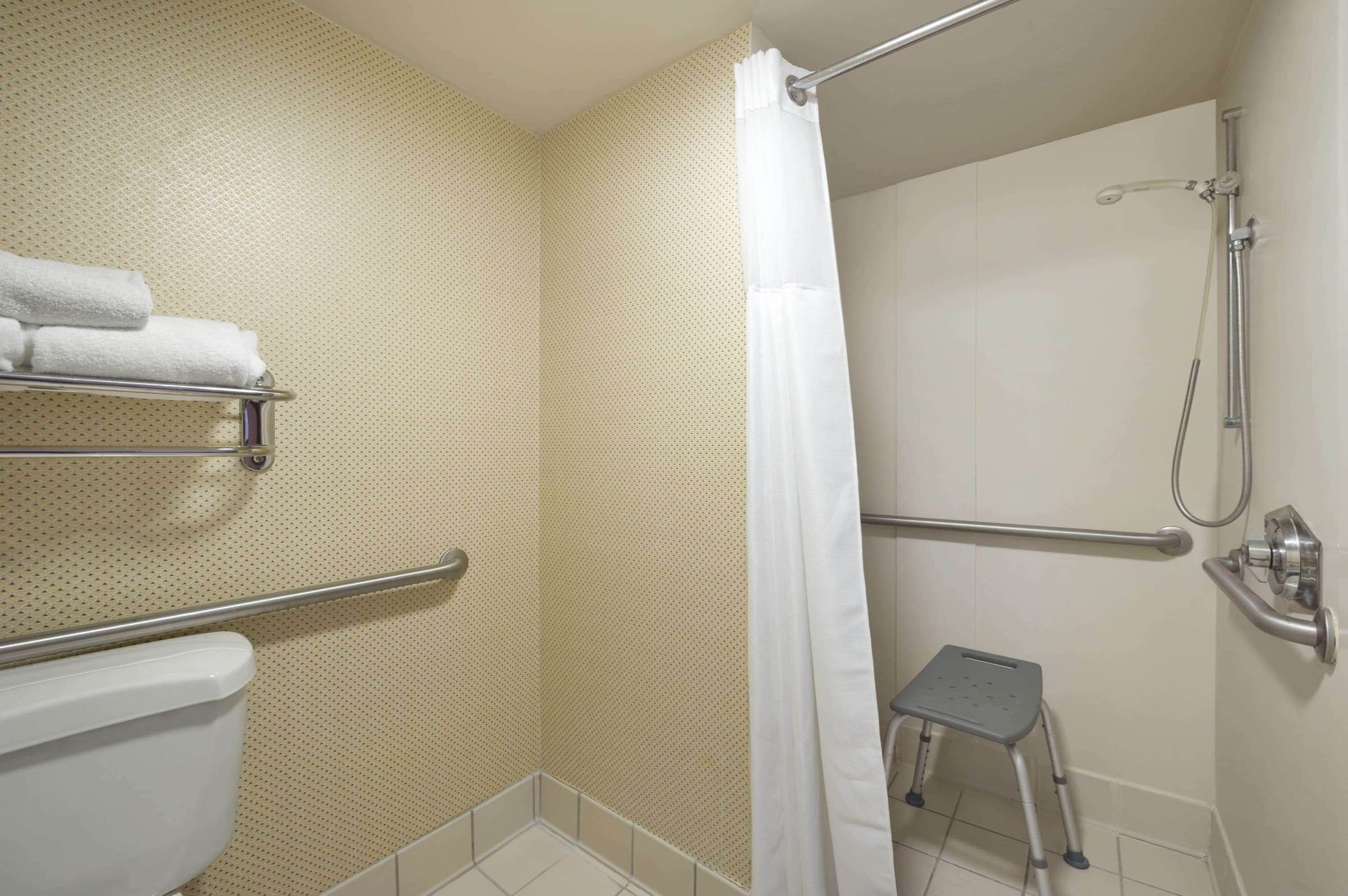 Un baño con inodoro, ducha con barra de apoyo, taburete y toallero.