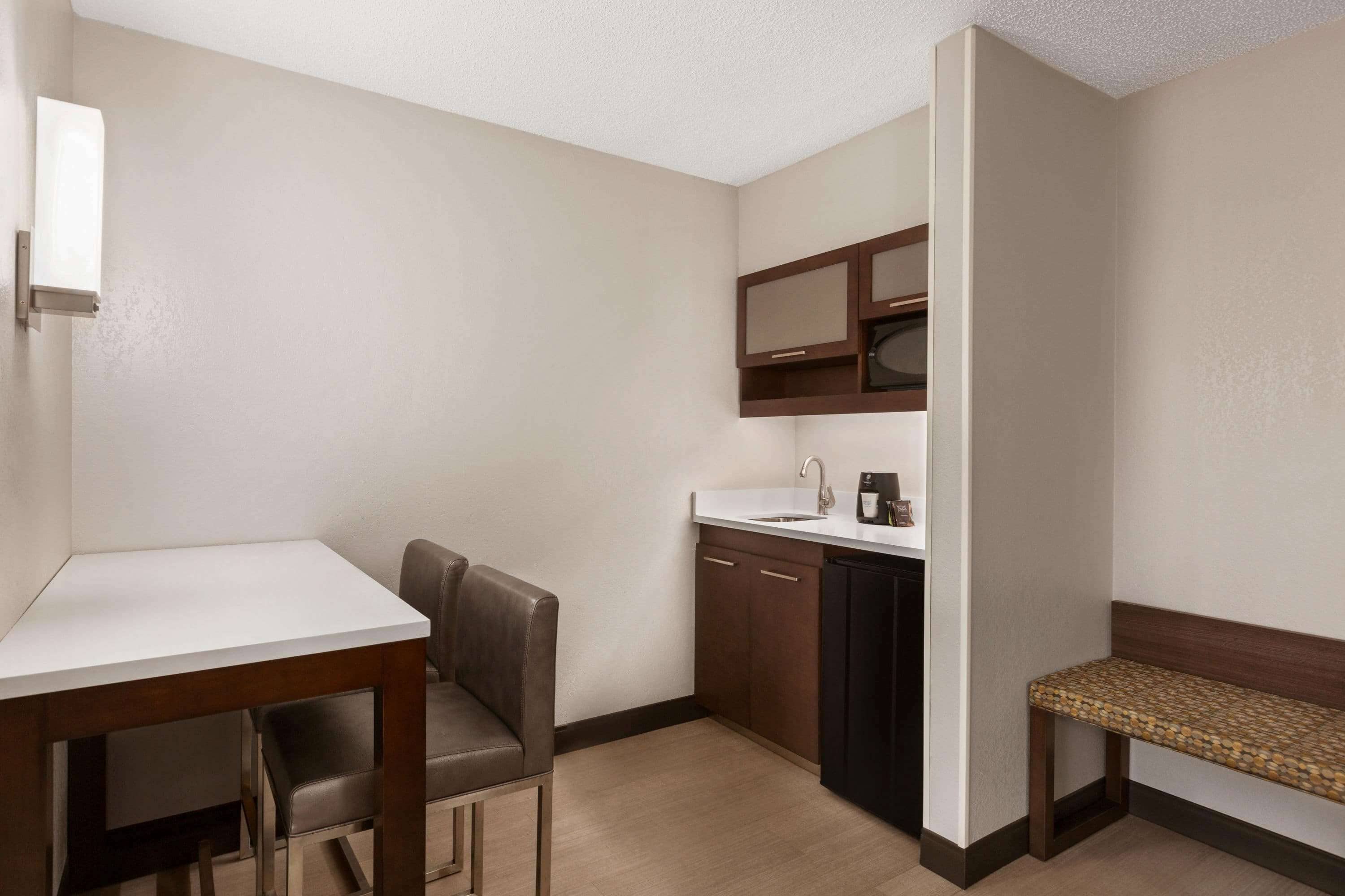 Una habitación de hotel compacta con kitchenette, una pequeña mesa de comedor, un banco y una sola silla.