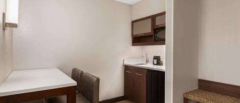 Una habitación de hotel compacta con kitchenette, una pequeña mesa de comedor, un banco y una sola silla.