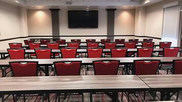 Een vergaderzaal met meerdere tafels en rode stoelen, voorzien van een groot scherm en een beamer.