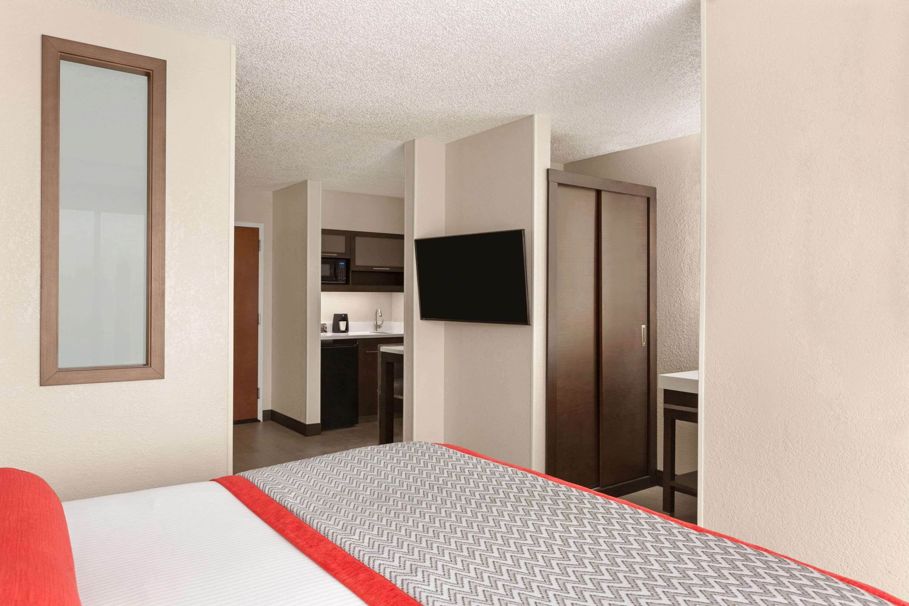 A hotel room with a bed, a television mounted on the wall, and a kitchenette in the background.