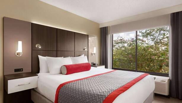 A modern hotel room with a large bed, a nightstand, a drawer, a wall-mounted light, and a window with a view of trees. - Ramada by Wyndham Suites Orlando Airport (Orlando)
