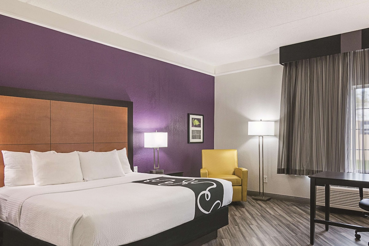 Ett hotellrum med en stor säng, en gul fåtölj, ett skrivbord och ett fönster med gardiner.