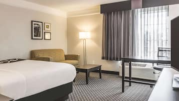 A hotel room with a large bed, a sofa, a desk, and a lamp. There are framed pictures on the wall and a window with curtains.