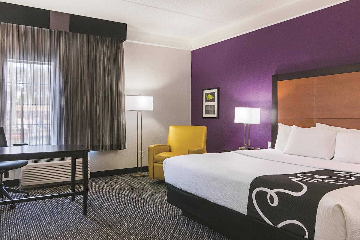 Ett hotellrum med en säng, ett skrivbord, en stol, en lampa och ett fönster med gardiner.