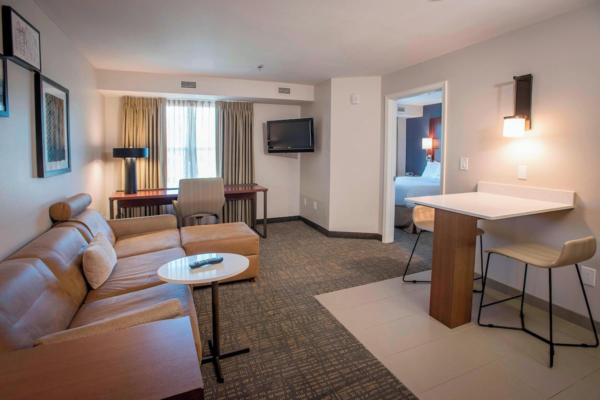A hotel room with a sofa, a small round table, a TV, a desk with a chair, and a bathroom visible through an open door.