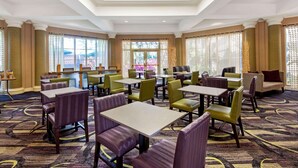 Un comedor de hotel con varias mesas y sillas, ventanas amplias con persianas y una alfombra estampada.