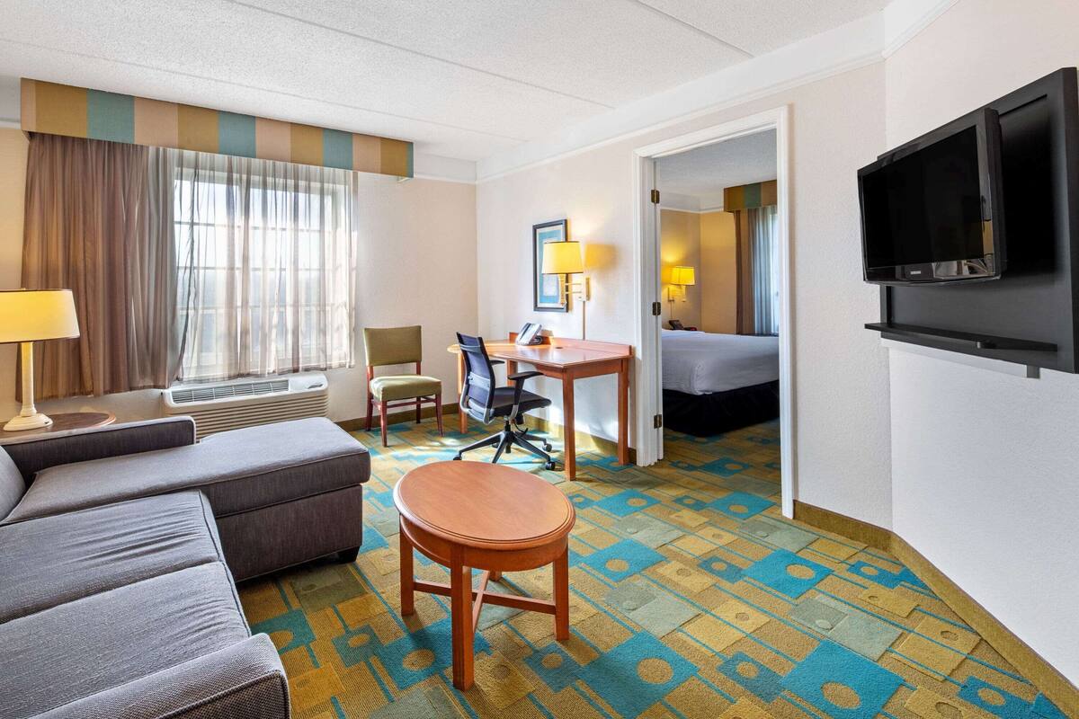 Una habitación de hotel con sofá, una mesa redonda de madera, un escritorio con silla, cama, televisión y una alfombra estampada.