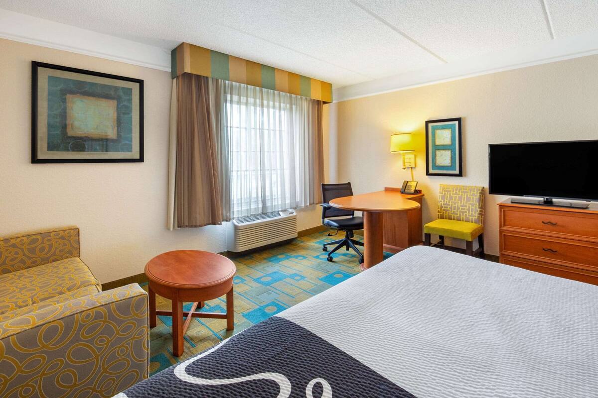Habitación de hotel con una cama grande, un escritorio, una silla, un taburete pequeño, un televisor y una ventana con cortinas.