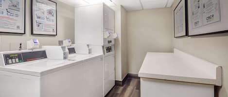 A laundry room with a washing machine, dryer, and ironing station. There are instructional posters on the walls.