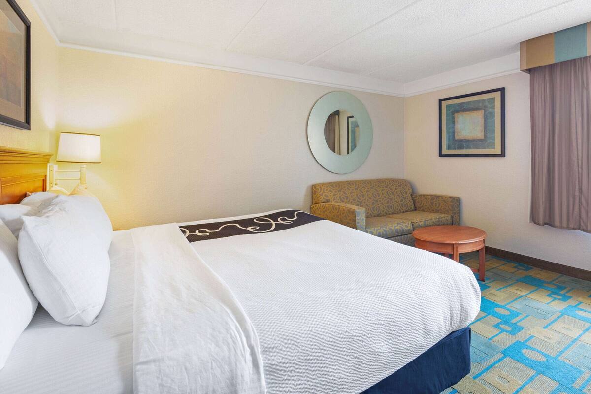 Una habitación de hotel con cama, una silla, una mesita, una lámpara y un espejo.