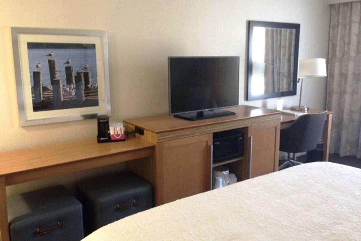 A hotel room with a bed, a desk with a television, a framed picture, a lamp, and a window with curtains.
