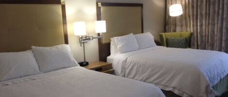 A hotel room with two beds, a nightstand with lamps, a chair, and a window with curtains.