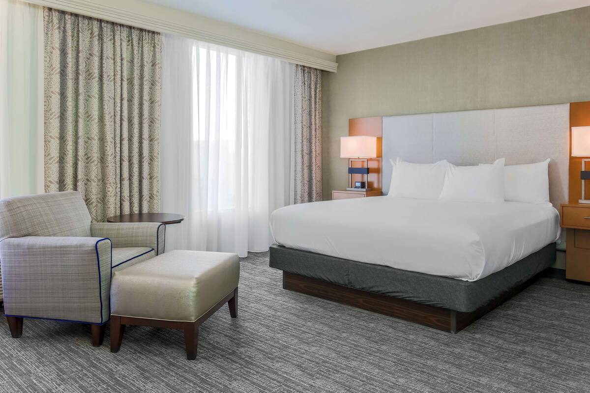 A hotel room with a large bed, a chair, an ottoman, a small table, and a lamp.